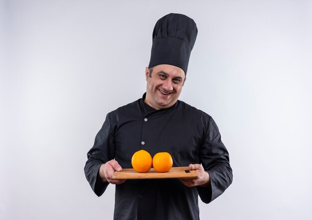 Smiling middle-aged male cook in chef uniform holding oranges on cutting board