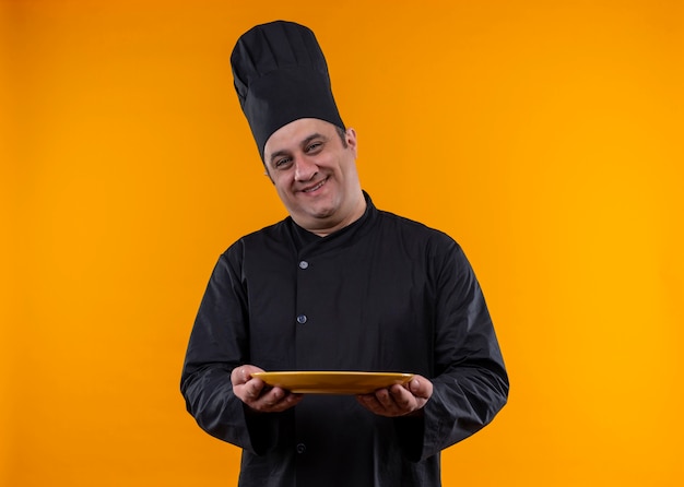 Smiling middle-aged male cook in chef uniform holding plate on yellow background with copy space