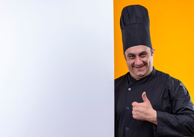 Smiling middle-aged male cook in chef uniform holding white wall his thumb up on yellow background with copy space