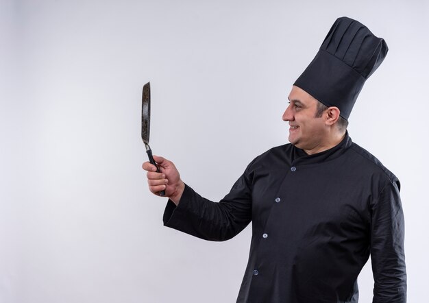 Smiling middle-aged male cook in chef uniform looking at frying pan in his hand with copy space