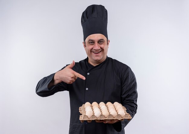 Smiling middle-aged male cook in chef uniform points to batch of eggs in his hand
