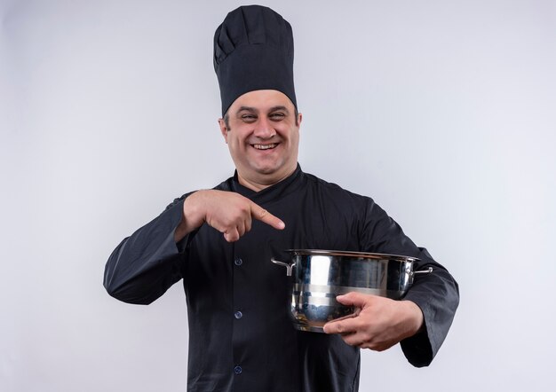 Smiling middle-aged male cook in chef uniform points at pot in her hand