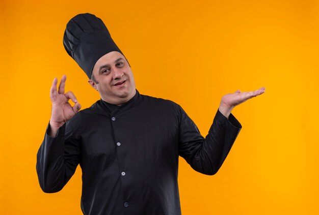 Smiling middle-aged male cook in chef uniform showing okey on yellow background with copy space