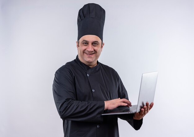 Smiling middle-aged male cook in chef uniform used laptop in his hand