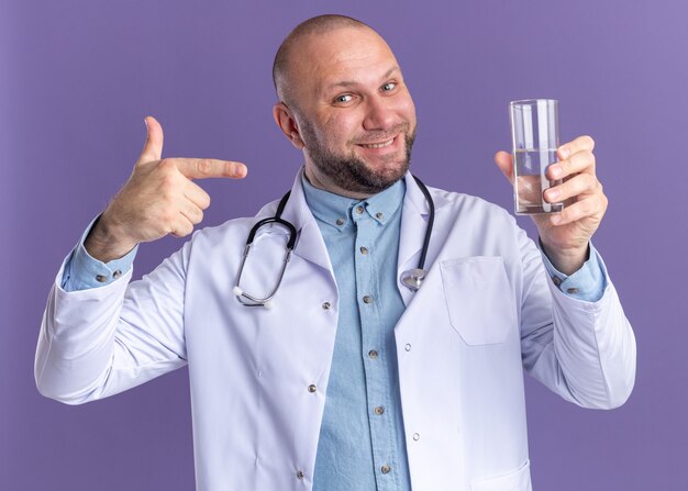 紫色の壁に隔離された水のガラスを保持し、指している医療ローブと聴診器を身に着けている中年男性医師の笑顔
