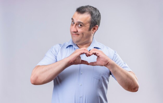 Smiling middle-aged man in blue vertical striped shirt showing heart sign with hands while standing on a white background
