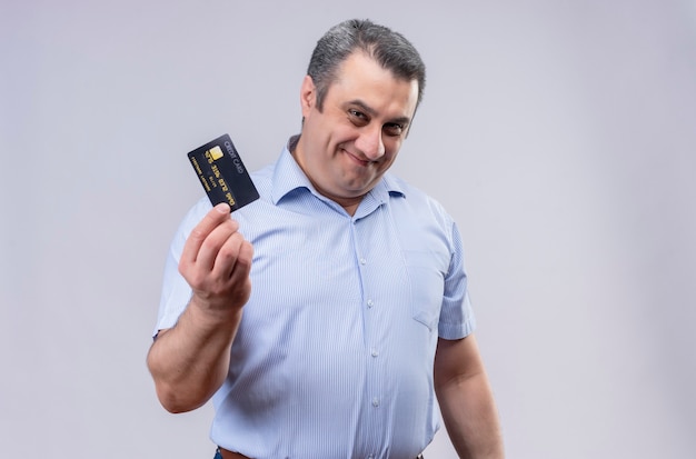 Smiling middle-aged man wearing blue vertical striped shirt holding credit card while standing on a white background