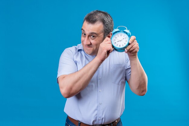 Smiling middle-aged man wearing blue vertical striped shirt listening to clock ticking sound holding blue alarm clock on a blue background