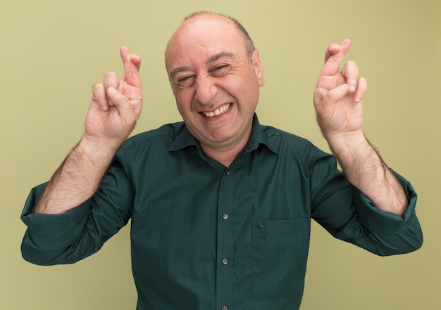 Улыбающийся мужчина средних лет в зеленой футболке, скрестив пальцы, изолирован на оливково-зеленой стене