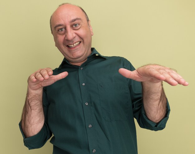 Smiling middle-aged man wearing green t-shirt holding out hands at camera isolated on olive green wall