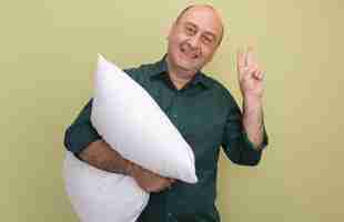 Free photo smiling middle-aged man wearing green t-shirt hugged pillow showing peace isolated on olive green wall