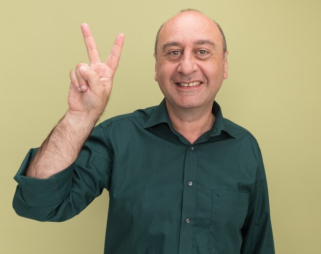 Smiling middle-aged man wearing green t-shirt showing peace gesture isolated on olive green wall