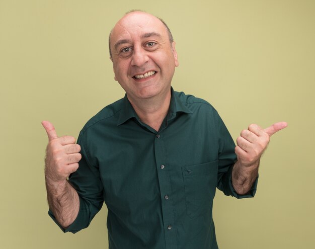 복사 공간 올리브 녹색 벽에 고립 된 측면에서 엄지 손가락과 포인트를 보여주는 녹색 티셔츠를 입고 웃는 중년 남자