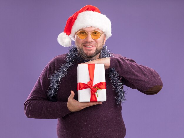 보라색 벽에 고립 된 선물 패키지를 들고 안경 목에 산타 모자와 반짝이 화환을 입고 웃는 중년 남자