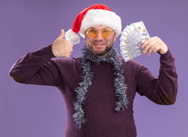 Smiling middle-aged man wearing santa hat and tinsel garland around neck with glasses holding money  showing thumb up isolated on purple wall Free Photo