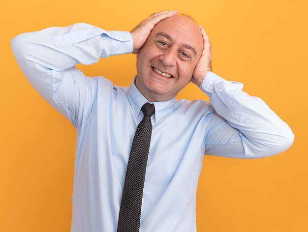 Улыбающийся мужчина средних лет в белой футболке с галстуком, положив руки на уши, изолирован на оранжевой стене