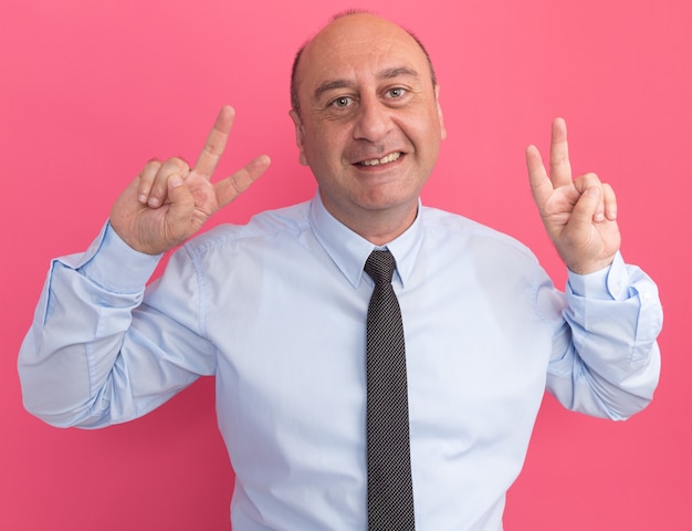 Foto gratuita sorridente uomo di mezza età che indossa la maglietta bianca con cravatta che mostra gesto di pace isolato sulla parete rosa