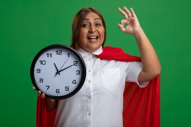 Smiling middle-aged superhero female holding wall clock and showing okay gesture isolated on green background