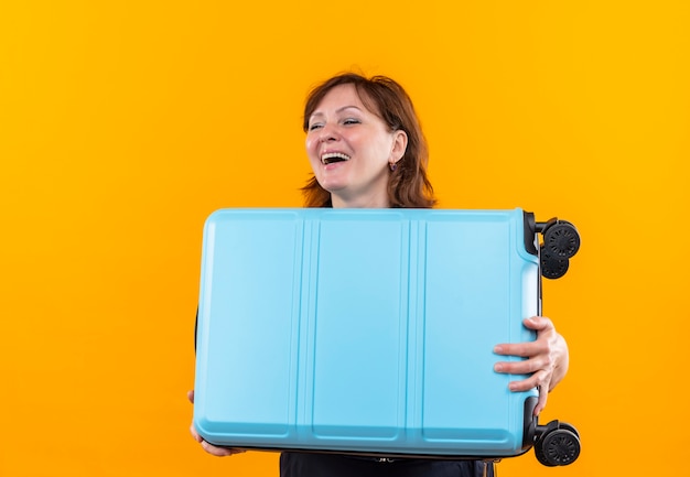 Smiling middle-aged traveler woman holding suitcase on isolated yellow