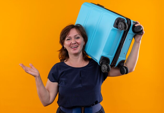 Smiling middle-aged traveler woman holding suitcase on shoulder and spread hand on isolated yellow wall