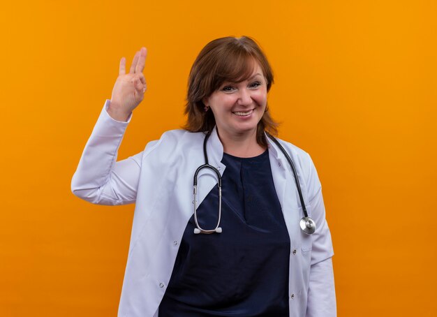 Улыбающаяся женщина-врач средних лет в медицинском халате и стетоскопе делает хорошо знак на изолированной оранжевой стене