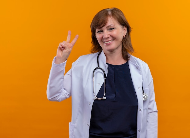 Smiling middle-aged woman doctor wearing medical robe and stethoscope doing peace sign on isolated orange wall with copy space