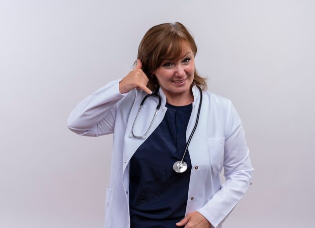 Smiling middle-aged woman doctor wearing medical robe and stethoscope doing phone gesture on isolated white wall