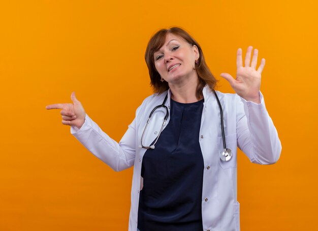 Smiling middle-aged woman doctor wearing medical robe and stethoscope pointing at left side and showing five on isolated orange wall