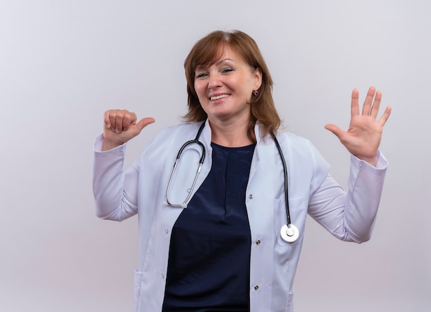 의료 가운과 청진기를 착용하고 자신을 손가락으로 가리키고 격리 된 흰 벽에 5를 보여주는 중년 여성 의사 미소