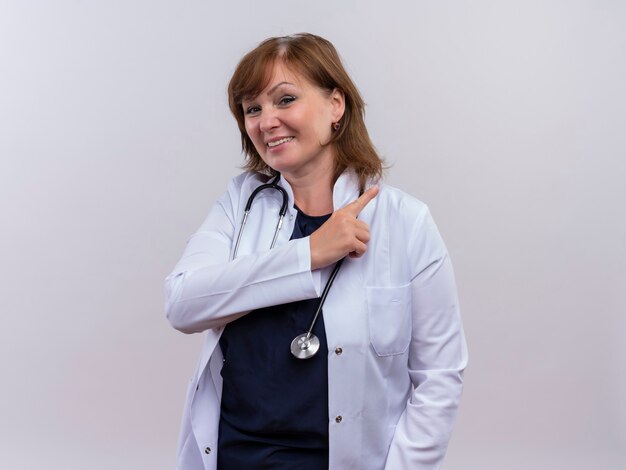 의료 가운 및 복사 공간이 격리 된 흰 벽에 오른쪽에 손가락으로 가리키는 청진기를 입고 웃는 중년 여성 의사
