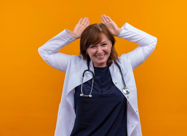 Smiling middle-aged woman doctor wearing medical robe and stethoscope putting hands over head on isolated orange wall