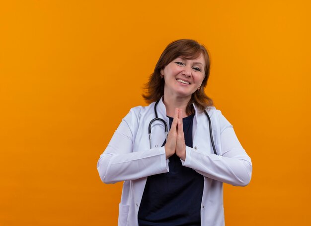 의료 가운과 청진기를 입고 웃는 중년 여성 의사가 복사 공간이 격리 된 주황색 벽에 손을 모으고 있습니다.