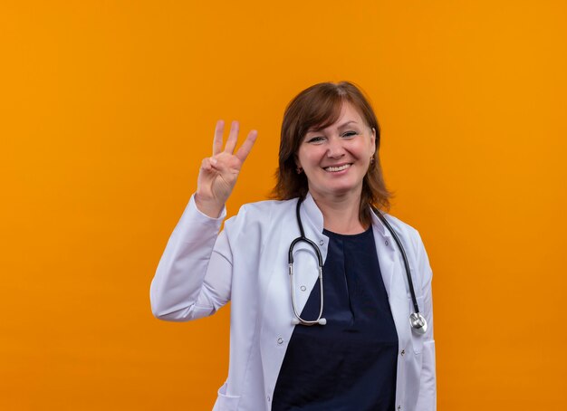 Smiling middle-aged woman doctor wearing medical robe and stethoscope showing three on isolated orange wall with copy space