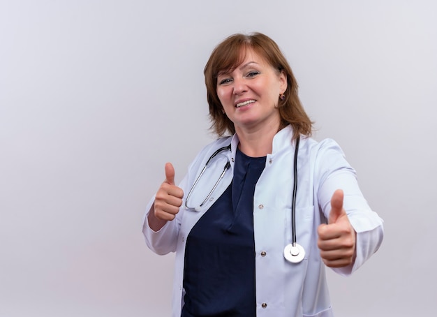 Smiling middle-aged woman doctor wearing medical robe and stethoscope showing thumbs up on isolated white wall with copy space