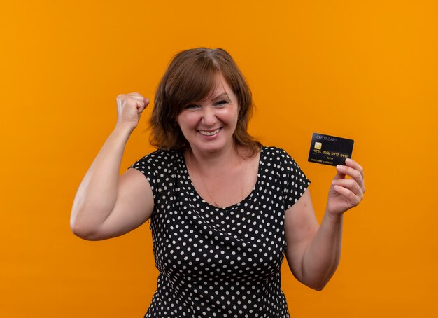Smiling middle-aged woman holding card and lifting her fist on isolated orange wall