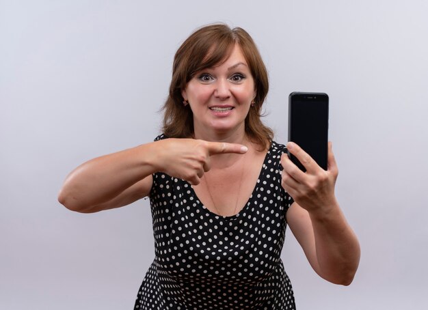 Smiling middle-aged woman holding mobile phone and pointing with finger at it on isolated white wall