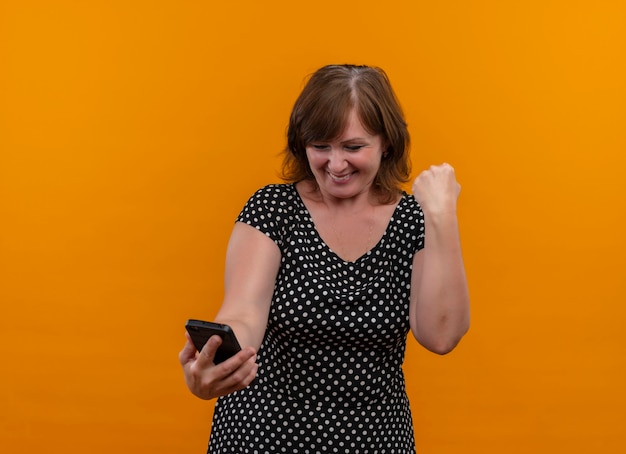Улыбающаяся женщина средних лет держит мобильный телефон с поднятым кулаком на изолированной оранжевой стене с копией пространства