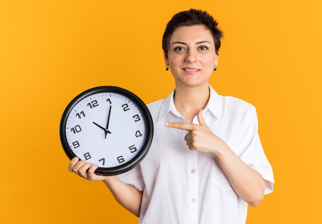 Smiling middle-aged woman holding and pointing at clock looking at camera 