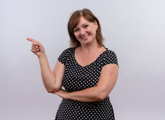Smiling middle-aged woman pointing with finger at left side on isolated white wall