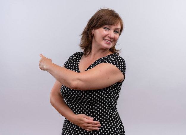 Улыбающаяся женщина средних лет показывает пальцем сзади и кладет руку на живот на изолированной белой стене