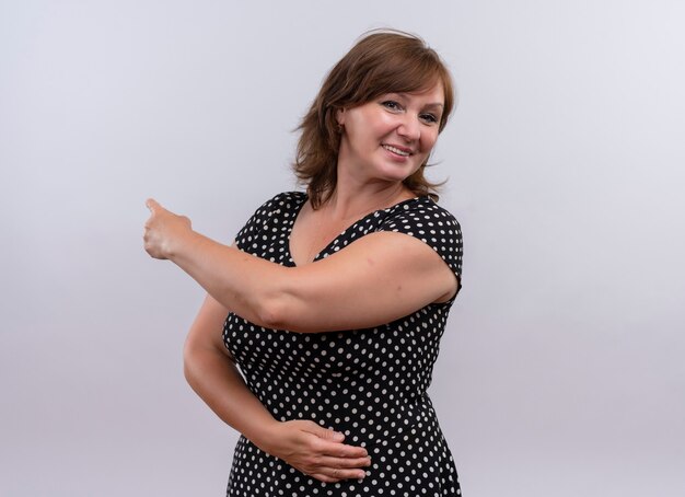 Smiling middle-aged woman pointing with finger behind and putting hand on belly on isolated white wall