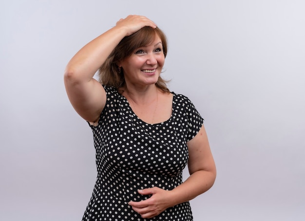Smiling middle-aged woman putting hands on head and belly on isolated white wall