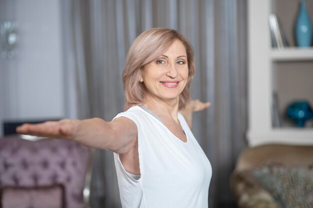 Smiling Middle Aged Woman Stretching Hands In Sides While Practising Yoga At Home. Yoga Practice Concept.