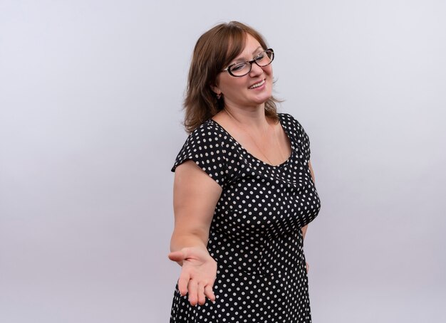 Smiling middle-aged woman wearing glasses and showing empty hand on isolated white wall