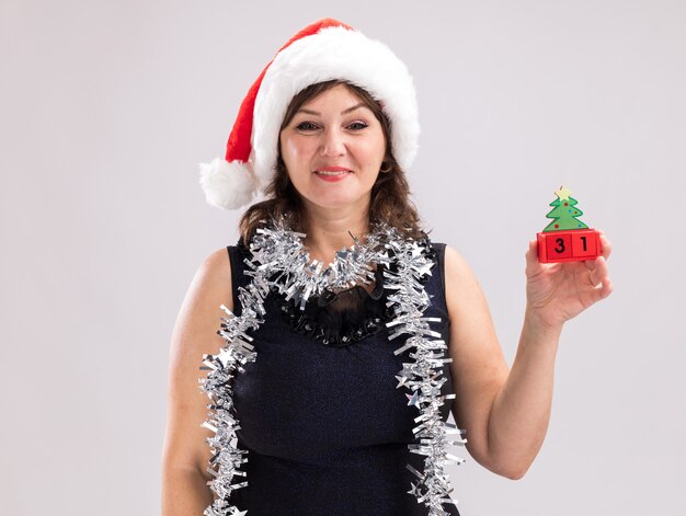 Smiling middle-aged woman wearing santa hat and tinsel garland around neck holding christmas tree toy with date looking at camera isolated on white background