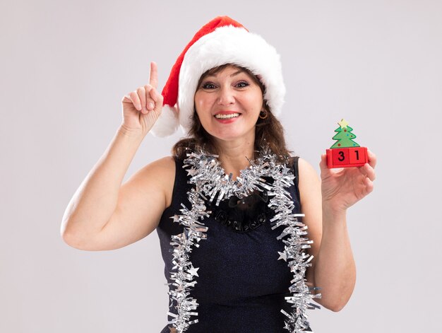 サンタの帽子と見掛け倒しの花輪を首に身に着けている中年の女性の笑顔は、白い背景で隔離のカメラを上向きに見ている日付とクリスマスツリーのおもちゃを保持しています