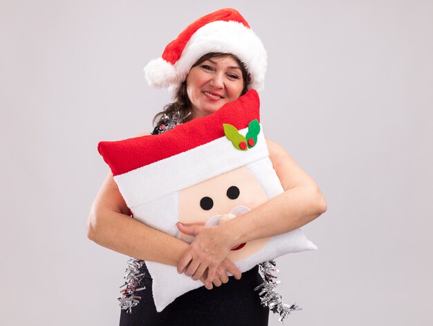 Smiling middle-aged woman wearing santa hat and tinsel garland around neck holding santa claus pillow looking at camera isolated on white background with copy space