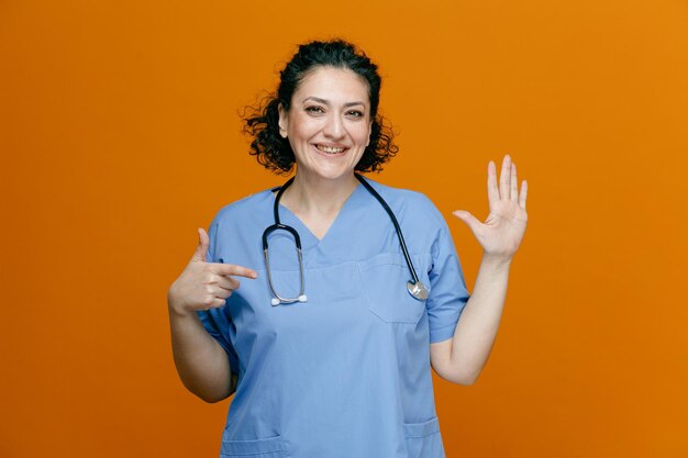 Улыбающаяся женщина-врач средних лет в униформе и со стетоскопом на шее смотрит в камеру, показывающую пятерых с рукой, указывающей на руку, изолированную на оранжевом фоне