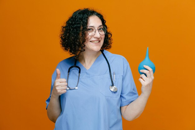 Smiling middleaged female doctor wearing uniform glasses and stethoscope around her neck showing enema looking at camera showing thumb up isolated on orange background