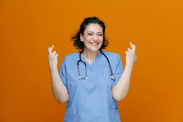 Smiling middleaged female doctor wearing uniform and stethoscope around her neck looking at camera making wish while crossing fingers isolated on orange background